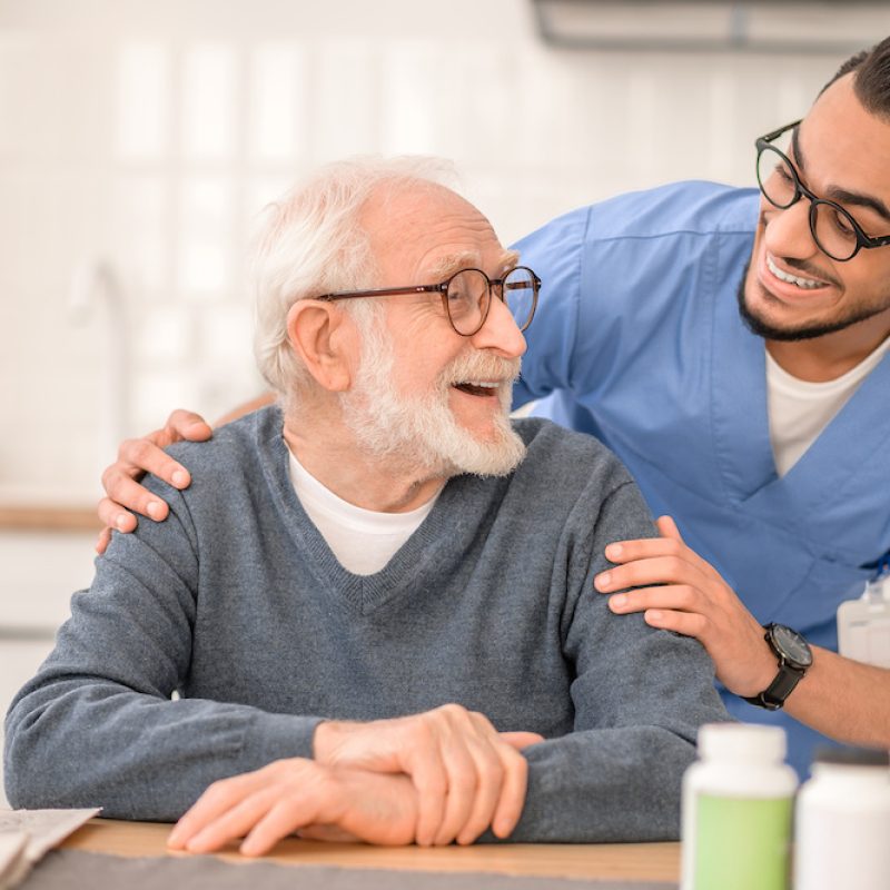 Smiling male nurse in uniform hugging a happy aged man seated at the kitchen table
